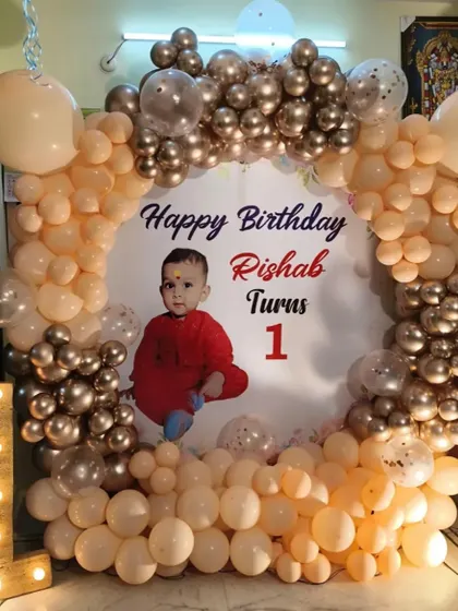 A first birthday decoration with a personalized photo backdrop. The setup for Rishab's first birthday includes a circular balloon frame and a light-up marquee number '1'.
