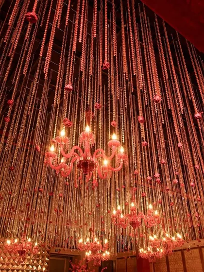 A breathtaking ceiling installation of hanging red floral strings and pink chandeliers, creating a dramatic and romantic ambiance for a Sangeet.