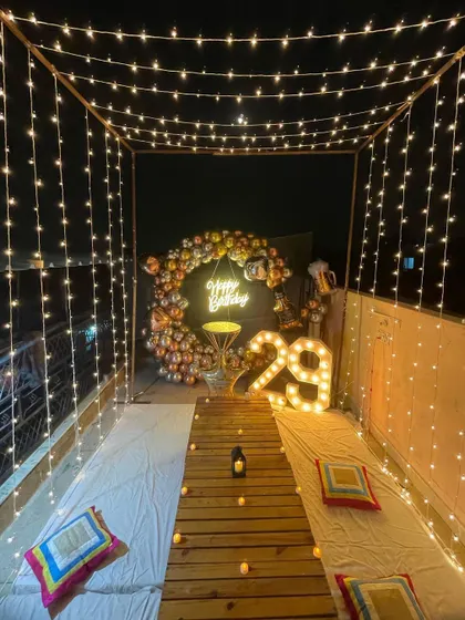 A stylish terrace birthday setup for a 29th birthday. The design includes a low pallet table, a canopy of lights, and a backdrop with a balloon arch and illuminated marquee numbers.