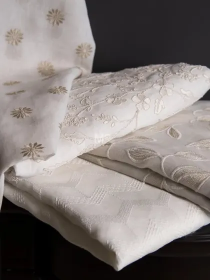 A stack of our sheer fabrics in off-white and ivory, featuring a variety of textures and patterns, including floral embroidery and geometric weaves, designed for effortless sophistication.