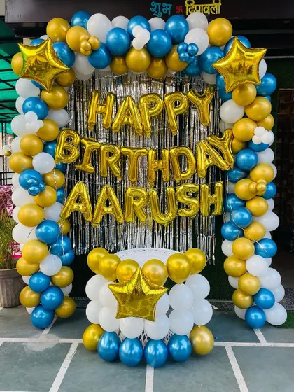 A bold blue, gold, and white balloon arch over a silver shimmer curtain. The "Happy Birthday" and name foil balloons add a personal touch.