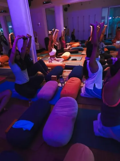 The studio transformed with ambient blue lighting for a special session. Students are seated, stretching their arms up to create space in the spine and shoulders.