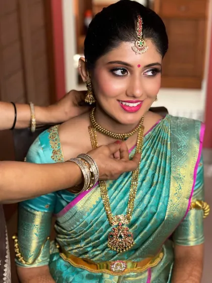 A candid moment as the final touches are put on the bride's jewelry. Her makeup is fresh and radiant, with a bright pink lip that complements her turquoise saree.