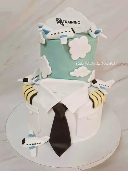 A two-tier corporate cake for an aviation training academy. The design combines a pilot's uniform on the bottom tier with a sky-themed top tier.