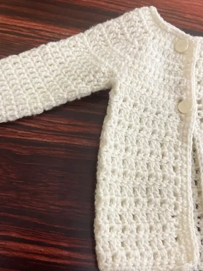 A detailed view of the sleeve and body of the off-white crochet baby cardigan, showcasing the uniform and neat stitch pattern.