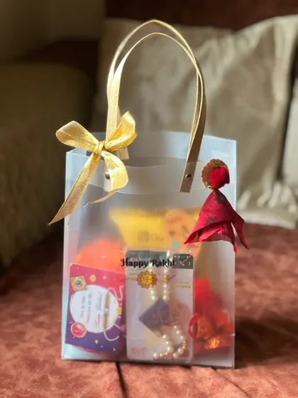 A chic and simple Rakhi gift presented in a transparent tote bag with a golden bow. This hamper includes a beautiful pearl rakhi and other small treats.
