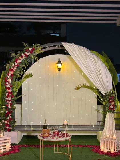 The full romantic proposal setup at night, featuring a floral arch, a table for two with champagne, and the ground covered in rose petals and candles.