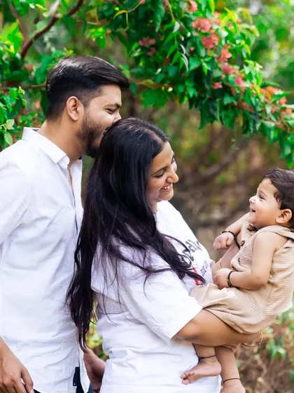 A candid moment of pure joy during an outdoor family photoshoot. The parents are all smiles as they play with their young child under the trees.