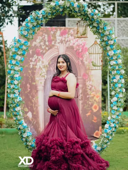 A stunning solo portrait of the mother-to-be in a maroon gown, framed by a beautiful floral arch.