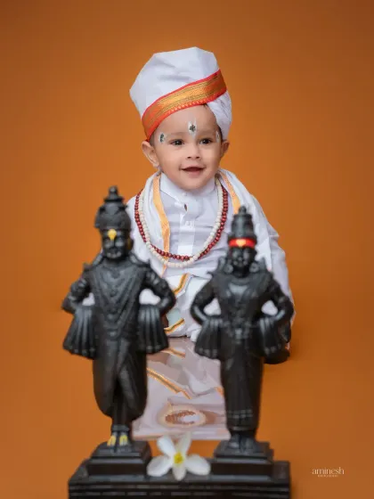 A smiling baby dressed in Varkari attire, posing with the idols of Vithoba and Rukmini. This is a simple yet powerful portrait against a solid orange background.