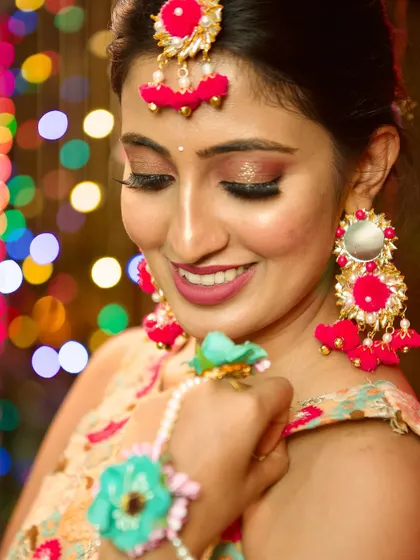 A joyful moment from the Mehendi event. The makeup features a warm, rosy blush and a matching lip color that brings out the festive colors of her outfit and the sparkling lights in the background.