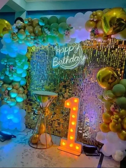 A first birthday party with a gold shimmer wall, a 'Happy Birthday' neon sign, and a '1' marquee light.