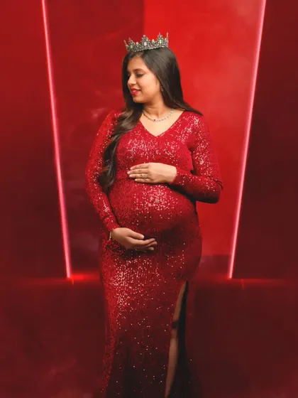 A regal and powerful portrait of a mom-to-be in a red sequin gown and crown, looking down at her bump with love.