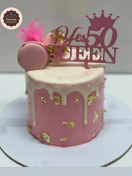 A chic and fun cake for a 50th birthday, with a "Yes 50 Queen" topper. The pink cake features a white chocolate drip and gold leaf, topped with a macaron for a touch of elegance.
