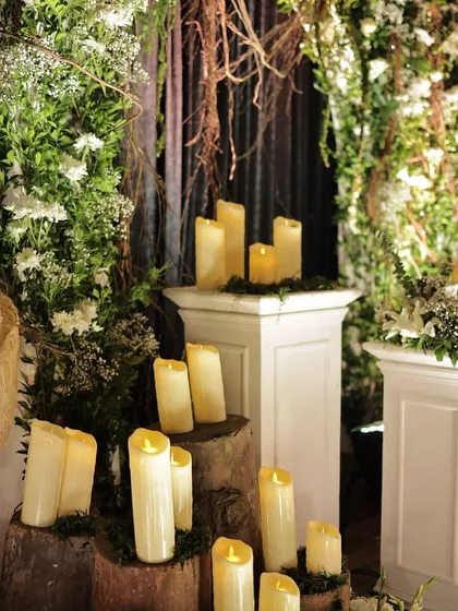 A close-up of the decor details from a rustic-themed event. We used pillar candles on wooden stumps and white pedestals, surrounded by lush greenery, to create a romantic, candlelit glow.