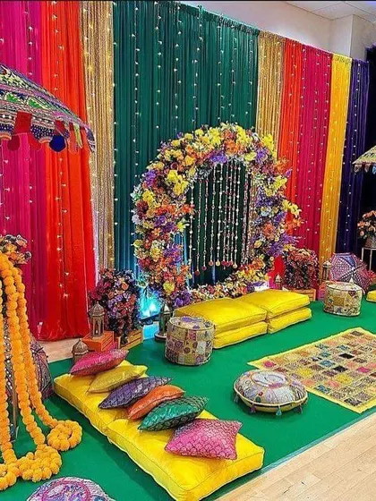 A vibrant and traditional Mehendi or Haldi seating area. The decor features a colorful backdrop of drapes, a floral ring, Rajasthani umbrellas, and low seating with bright cushions for a festive feel.