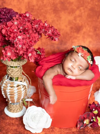 This newborn is sleeping in a red bucket, surrounded by red and white flowers.