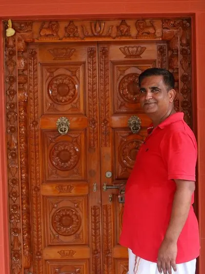 The entrance to the Ram's Anugraham residence features an intricately carved wooden door set against a bold, red-orange Porotherm brick facade, creating a warm and traditional welcome.