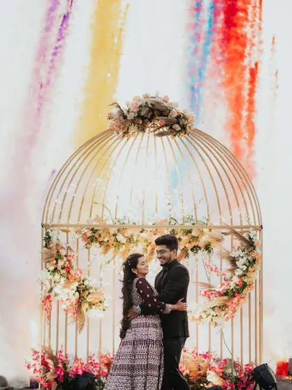 A romantic portrait of a couple at their beachside engagement, framed by a golden birdcage and colorful smoke.