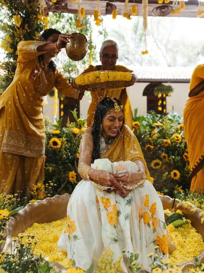 A beautiful moment during the Haldi ritual, where the bride is lovingly showered with flower petals and turmeric water by her family. The decor is filled with bright yellow flowers, enhancing the festive spirit.