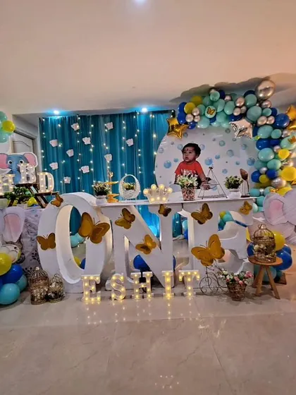 A cute baby elephant theme for Eshit Gowda's first birthday. The decor includes a "ONE" table, elephant cutouts, and a blue, yellow, and green color scheme for a playful celebration.