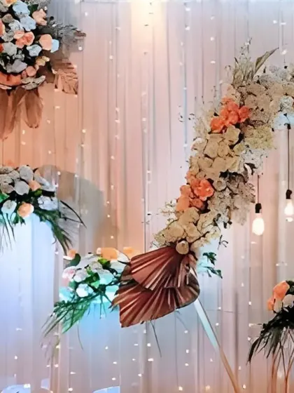 An asymmetrical floral arch in peach and white tones creates a soft and romantic focal point, complemented by hanging Edison bulbs for a warm, ambient light.