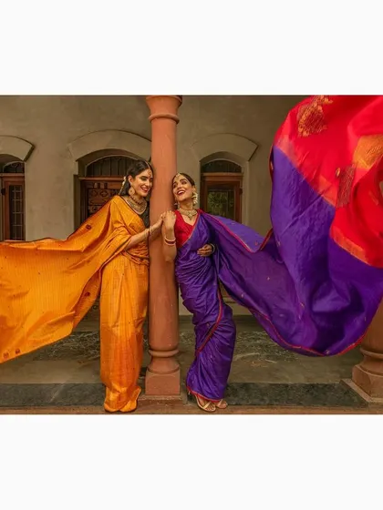 Two models showcasing vibrant silk sarees. The wind-blown fabric creates a sense of dynamic energy and highlights the beautiful drape and color of the garments.