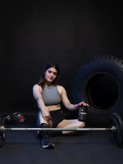 A female model resting on the gym floor with a barbell, holding a BCAA supplement. This shot captures a moment of rest and recovery.