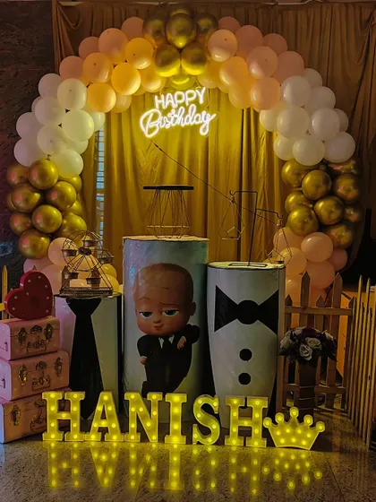 A stylish Boss Baby theme for Hanish, featuring custom-printed pedestals with a suit and tie design, a golden curtain backdrop, and marquee letters spelling out his name.