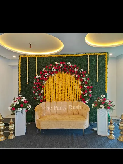 A classic floral backdrop for Varsha and Ajay's baby shower. The green hedge wall is adorned with a semi-arch of red, white, and yellow flowers, creating a vibrant and traditional setting for the celebration.