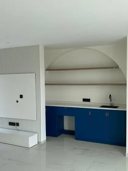 A bright and modern family lounge and pantry area. The custom-built pantry features deep blue cabinets, a white countertop, and open wooden shelves set within a stylish arch.
