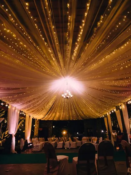 A dreamy wedding setup under a canopy of fairy lights. The draped fabric and central chandelier create a magical and romantic atmosphere for an evening event.