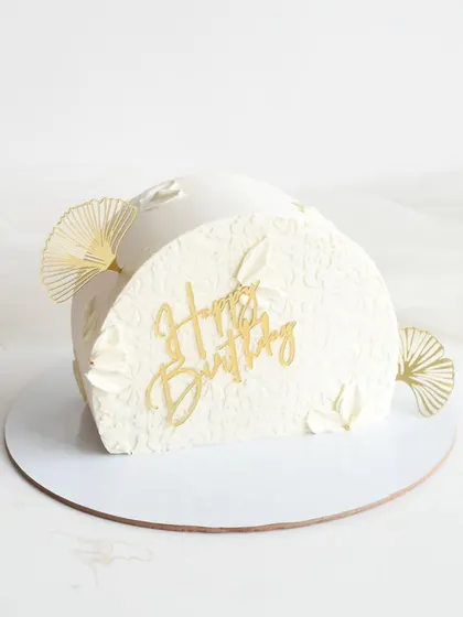 A modern and elegant half-birthday cake. The unique semi-circular shape is finished with a white textured frosting and accented with gold fans and a "Happy Birthday" topper.