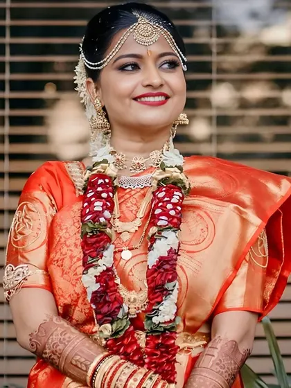 A radiant bride ready for her wedding ceremony. Her makeup features a classic red lip and defined eyes, perfectly complementing her red and gold saree and the traditional flower garlands.