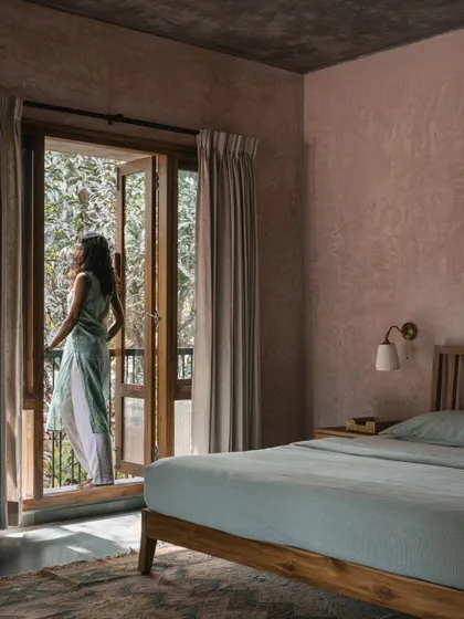 A bedroom featuring soft, pink-hued lime-washed walls and a simple wooden bed frame. French doors open onto a private balcony, letting in ample light and fresh air, and offering a view of the surrounding greenery.