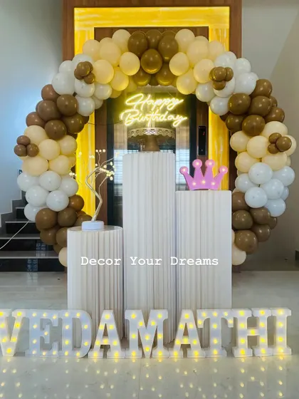A sophisticated birthday party with a brown and beige balloon arch. The setup includes marquee letters and a light-up crown.