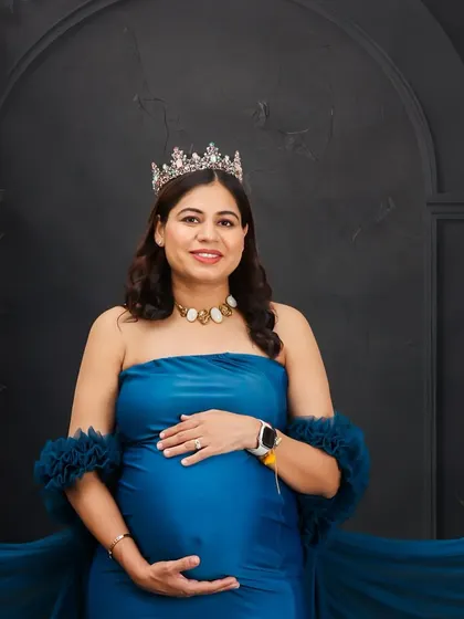 A classic portrait of a mother-to-be in a blue gown and crown, cradling her bump. The dark, textured studio backdrop adds depth and elegance to the shot.