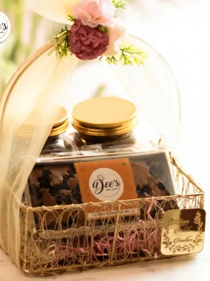 A golden wire basket hamper, elegantly wrapped for Raksha Bandhan. It contains jars of my signature baked goods and is adorned with a floral arrangement on the handle.