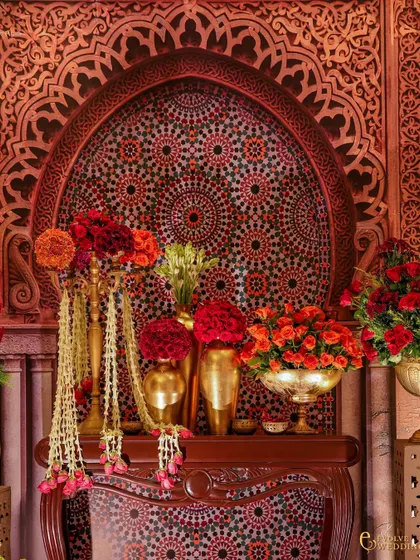 An ornate console table featuring a mosaic arch backdrop inspired by Indo-Islamic art. The arrangement includes brass vases with red and orange flowers and cascading jasmine garlands, embodying the aesthetic of abundance and grace.