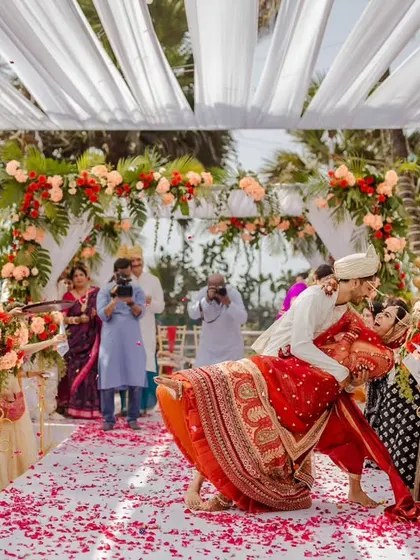 A playful and romantic moment from the wedding ceremony. The groom lifts his bride, surrounded by celebrating guests, on an aisle covered in rose petals. The floral arch and white canopy create a beautiful backdrop.