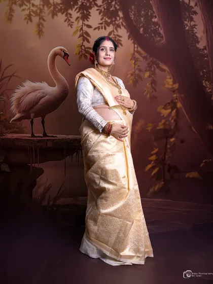 A graceful portrait of a mom-to-be in a traditional cream and gold saree, set against a painted swan backdrop for an artistic and cultural touch.