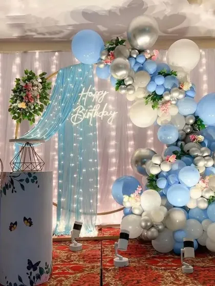 A dreamy blue and silver theme perfect for a serene anniversary celebration. This backdrop uses a sheer blue curtain with fairy lights, complemented by a balloon arch in shades of blue, white, and silver chrome.