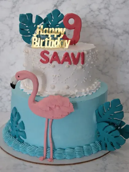 A stylish two-tier flamingo cake for a 9th birthday. The design features a handmade pink flamingo, tropical leaves, and a modern gold "Happy Birthday" topper.