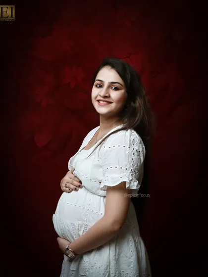 A radiant smile from the mom-to-be in a lovely white eyelet dress against a rich red studio backdrop.