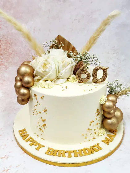We celebrate a 60th birthday in style with this chic white and gold floral cake. Fresh white roses and dried pampas grass create a modern botanical arrangement, while gold spheres and lettering add a touch of luxury to the pristine ivory cake.