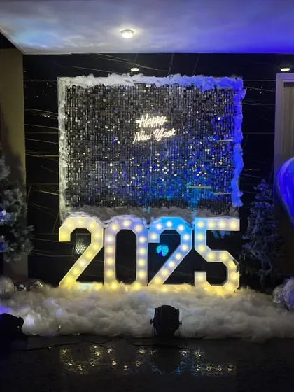 A New Year's Eve party decoration for 2025. The setup features a silver shimmer wall, large marquee numbers, and a winter theme with artificial snow and Christmas trees.
