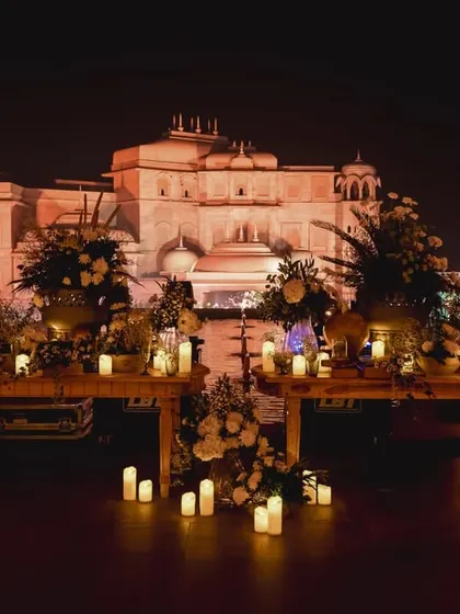 A beautiful welcome dinner setup with the Fairmont Jaipur palace lit up in the background. The long wooden tables are decorated with candles and floral urns for a rustic-regal feel.