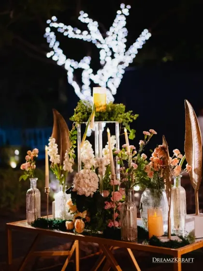 An elegant table setting for a reception, featuring a mix of fresh florals, taper candles, and gold accents. The lit-up tree in the background adds a layer of depth and magic to the outdoor setting.