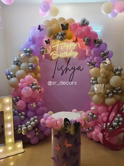 A whimsical butterfly theme for Tishya's first birthday, featuring a pastel balloon arch with silver accents, large butterfly cutouts, and a custom-decorated cake stand.