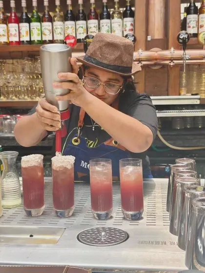 A female bartender expertly prepares a row of tall cocktails, adding a foam topping to each one with precision.